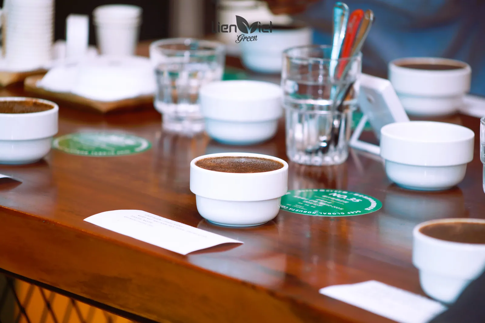 Cận cảnh chén cupping sứ trắng, cốc nước và thìa đặt trên bàn gỗ, phiếu ghi mẫu phía trước, hậu cảnh xoá phông
