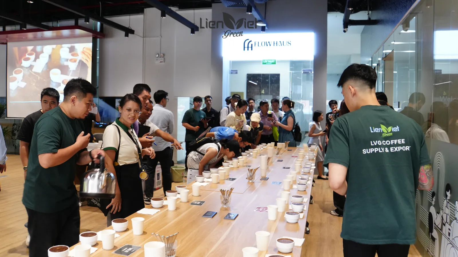 Nhân viên Liên Việt Green mặc áo LVG Coffee Supply & Export điều phối khách thử mẫu bên bàn cupping dài, tại sự kiện The New Crop – Trải nghiệm mùa vụ mới