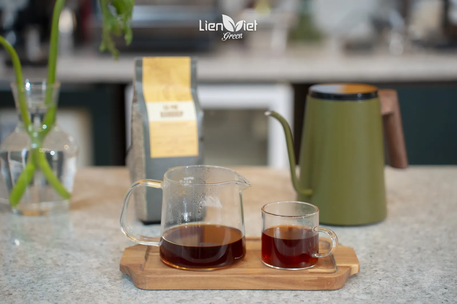 Pour-over carafe and a cup of black coffee on a wooden tray next to a kettle and a coffee bag
