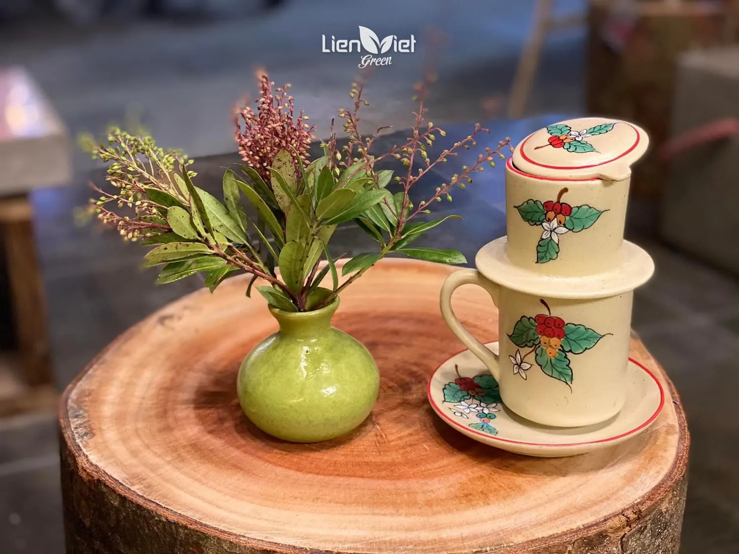 Phin gốm vẽ tay hoạ tiết cà phê trên cốc và đĩa, đặt trên mặt gỗ tròn cạnh bình hoa nhỏ
