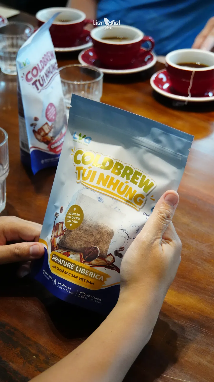Hand holding a Liberica cold brew steeped coffee bag beside coffee cups